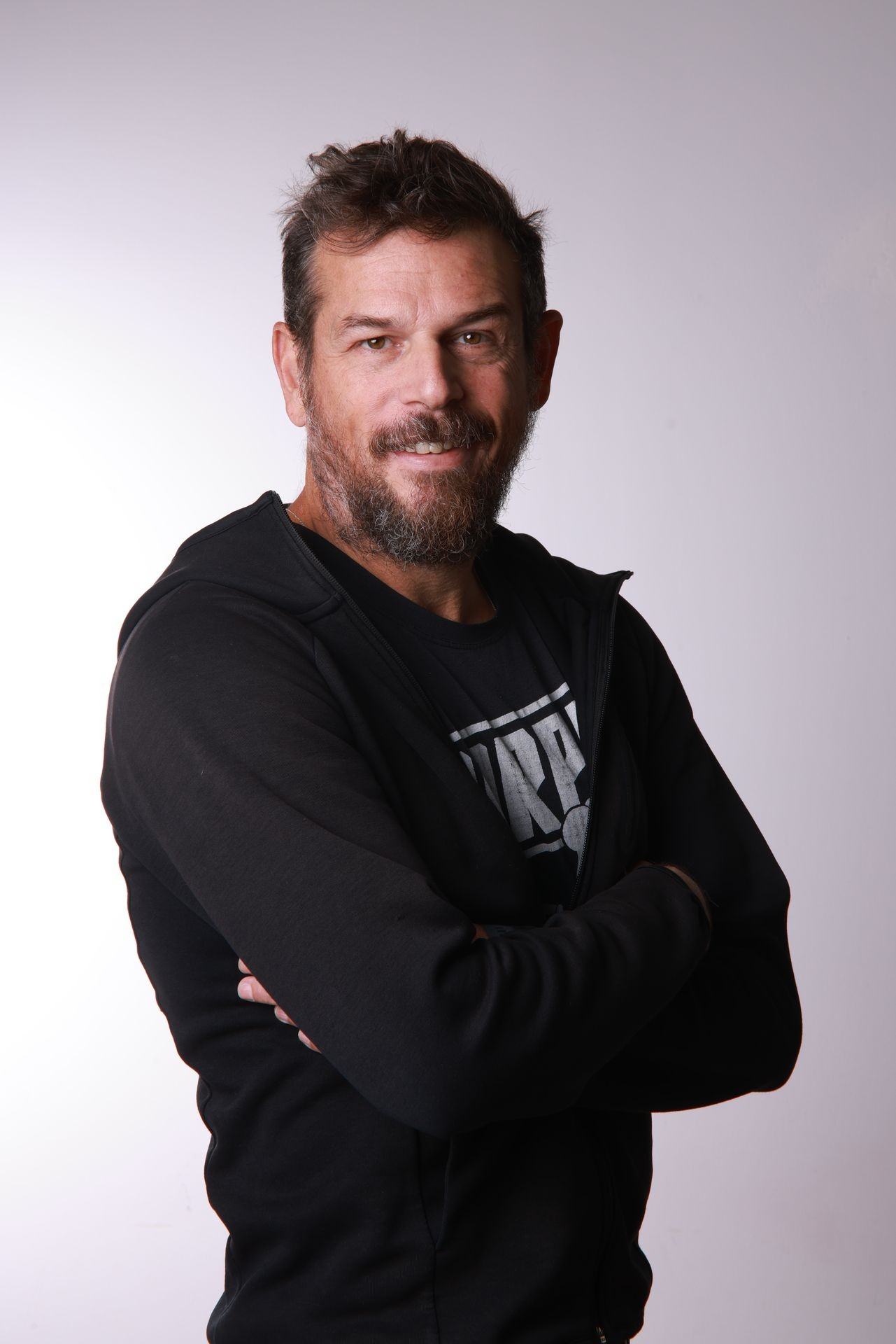 Bearded man in black jacket smiling with arms crossed against a plain background.